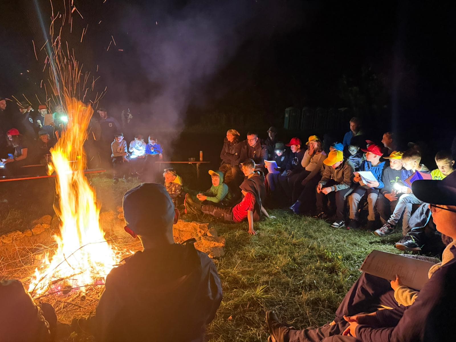Kinder und Betreuer*innen sitzen nachts im Kreis um ein großes Lagerfeuer
